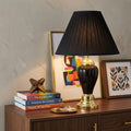 27" Black and Gold Ceramic Urn Table Lamp With Black Empire Shade displayed on a dark wood tabletop in a furnished interior against a tan wall.