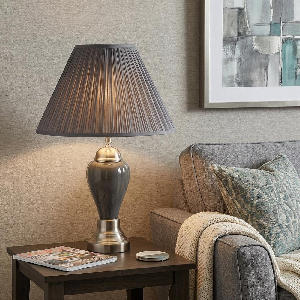 27" Gray and Silver Metal Urn Table Lamp With Gray Empire Shade displayed on a dark wood tabletop in a furnished interior against a tan wall.