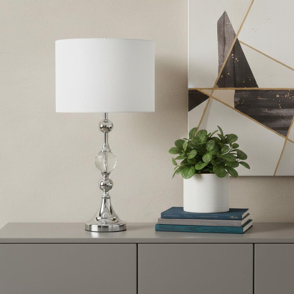 30" Silver Metal Bedside Table Lamp With White Classic Drum Shade displayed on a tabletop in a furnished interior against a cream wall.