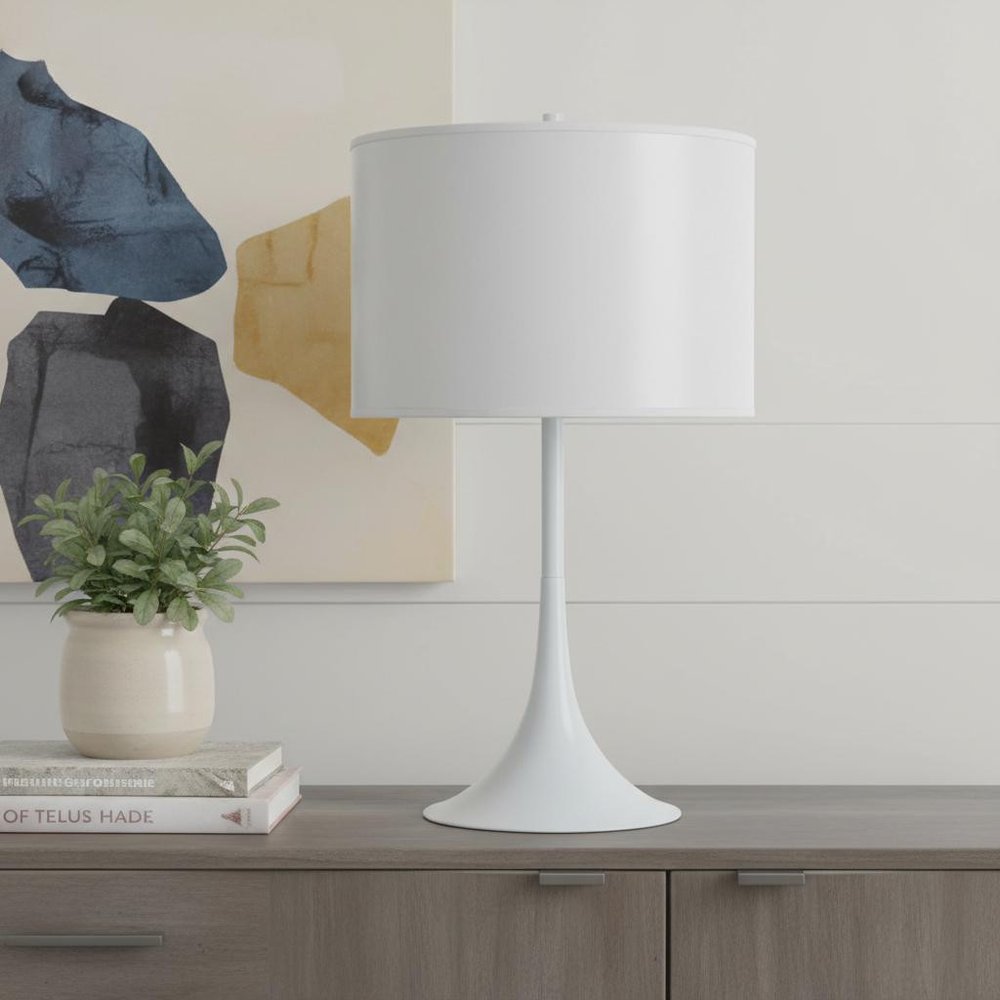 25" White Metal Table Lamp With White Classic Drum Shade displayed on a wood tabletop in a furnished interior against an off-white wall.