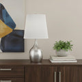 25" Silver Metal Gourd Table Lamp With White Tapered Drum Shade displayed on a dark wood tabletop in a furnished interior against a cream wall.