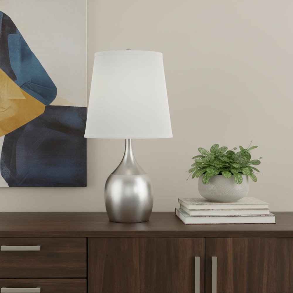 25" Silver Metal Gourd Table Lamp With White Tapered Drum Shade displayed on a dark wood tabletop in a furnished interior against a cream wall.