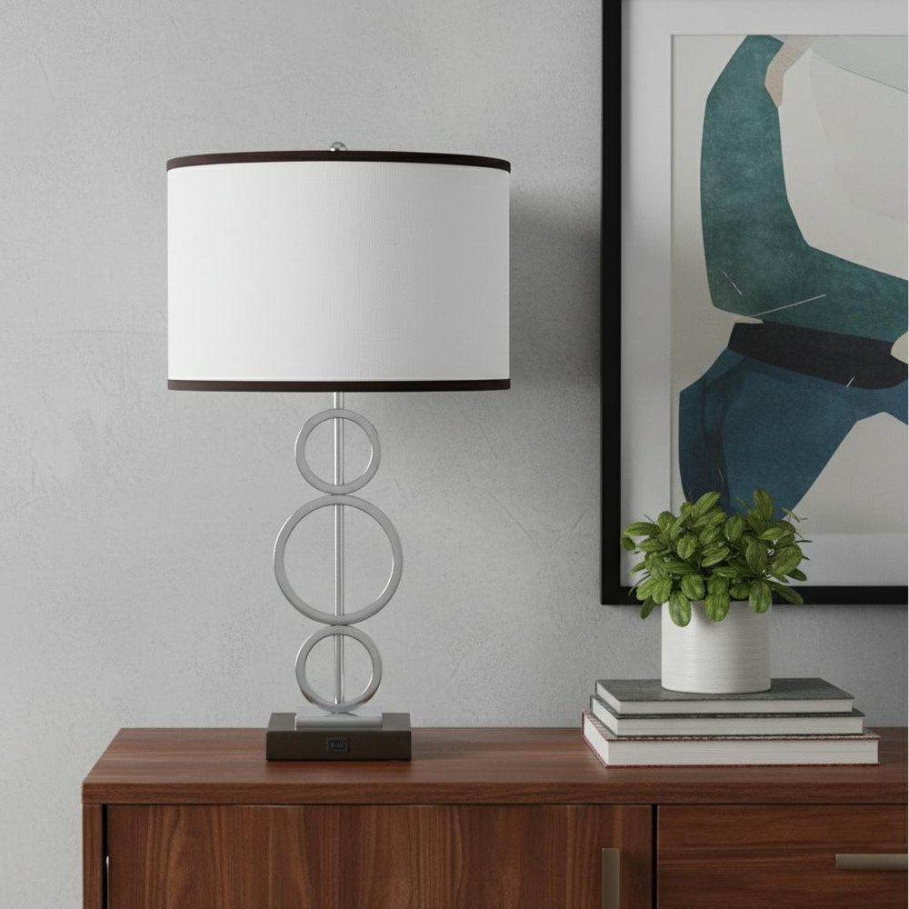 29" Silver Metal Bedside Geo Table Lamp With White and Black Trim Classic Drum Shade displayed on a dark wood tabletop in a furnished interior against a light gray wall.