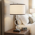 29" Silver Metal Bedside Geo Table Lamp With White and Black Trim Classic Drum Shade displayed on a dark wood tabletop in a furnished interior against a cream wall.