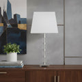 25" Crystal Geo Cubed Table Lamp With White Sharp Corner Square Tapered Shade displayed on a dark wood tabletop in a furnished interior against a gray wall.