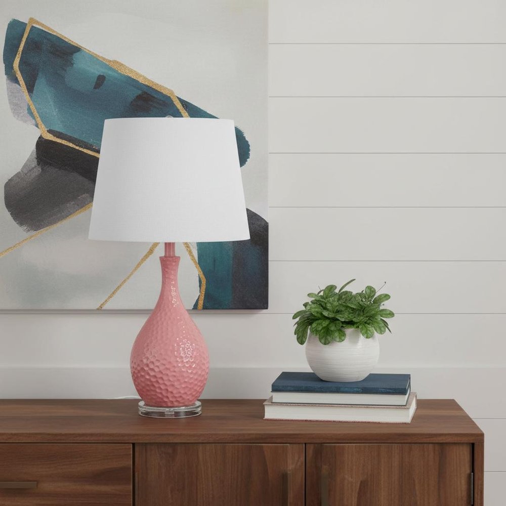28" Pink Hammered Urn Table Lamp With White Tapered Drum Shade displayed on a dark wood tabletop in a furnished interior against a light gray wall.