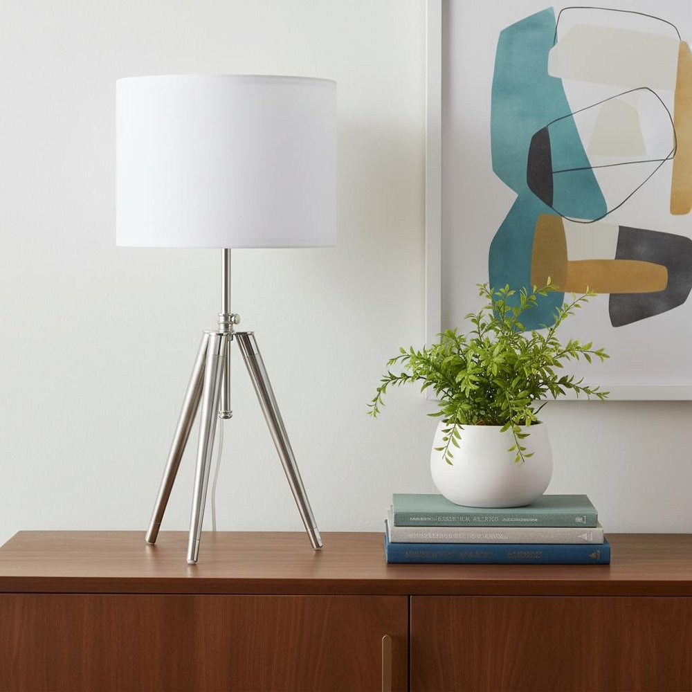 34" Silver Metal Adjustable Tripod Table Lamp With White Round Shade displayed on a dark wood tabletop in a furnished interior against an off-white wall.