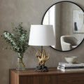 26" Natural And Brown Antlers Table Lamp With White Shade displayed on a dark wood tabletop in a furnished interior against a gray wall.