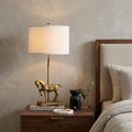 30" Gold Stallion Horse Table Lamp With White Shade displayed on a dark wood tabletop in a furnished interior against a tan wall.