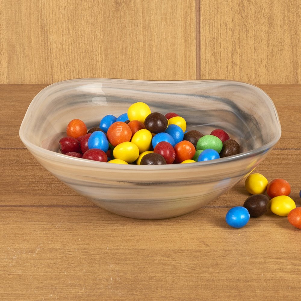 A square alabaster glass bowl filled with colorful M&M candies sits on a wooden surface.