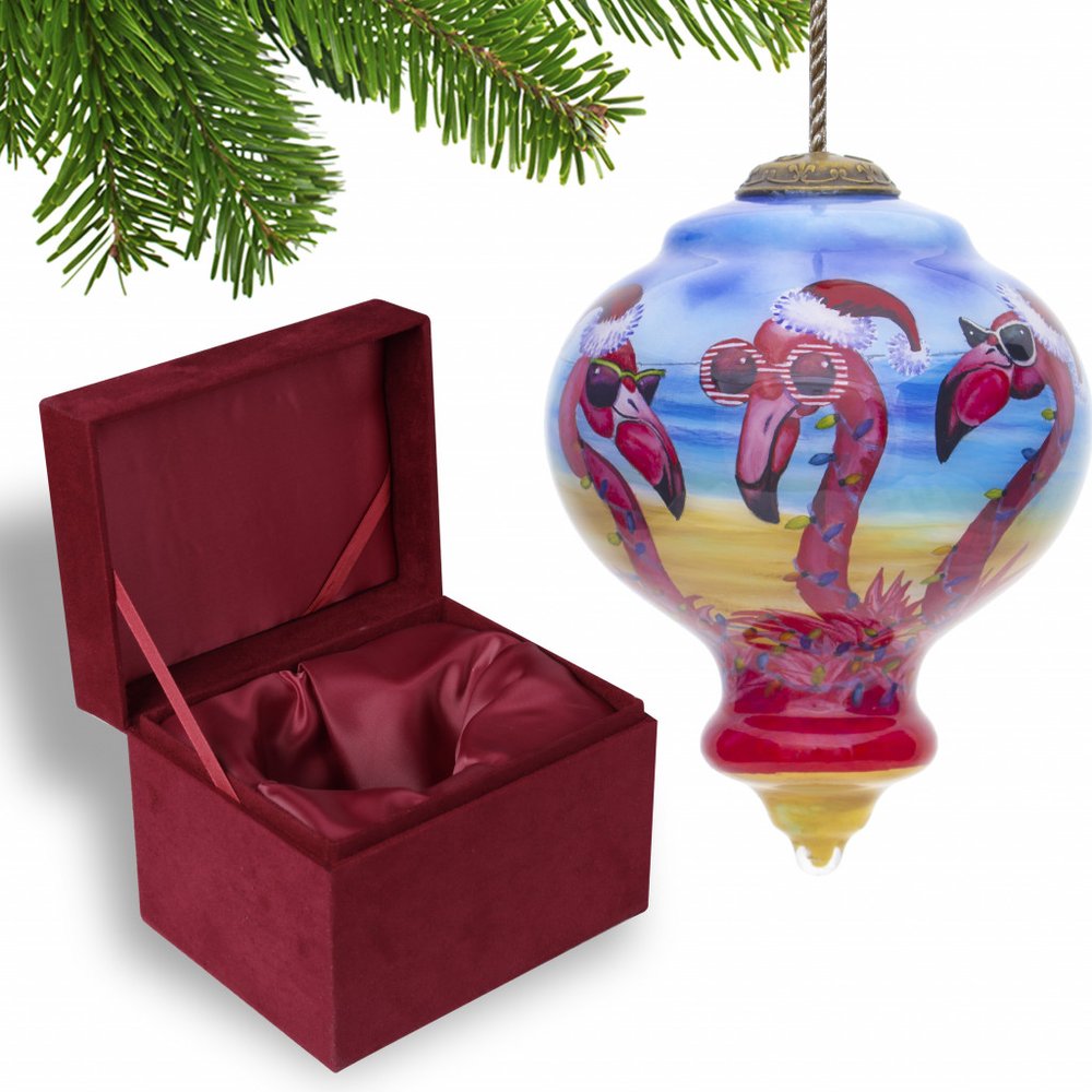 Red velvet jewelry box with satin lining next to a glass finial ornament with three flamingos wearing Santa hats and sunglasses on a beach.