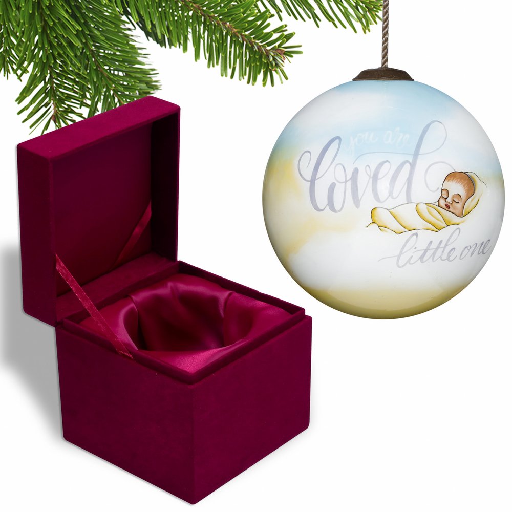 A round glass Christmas ornament with a baby and text "you are loved little one" sits next to an open velvet box.
