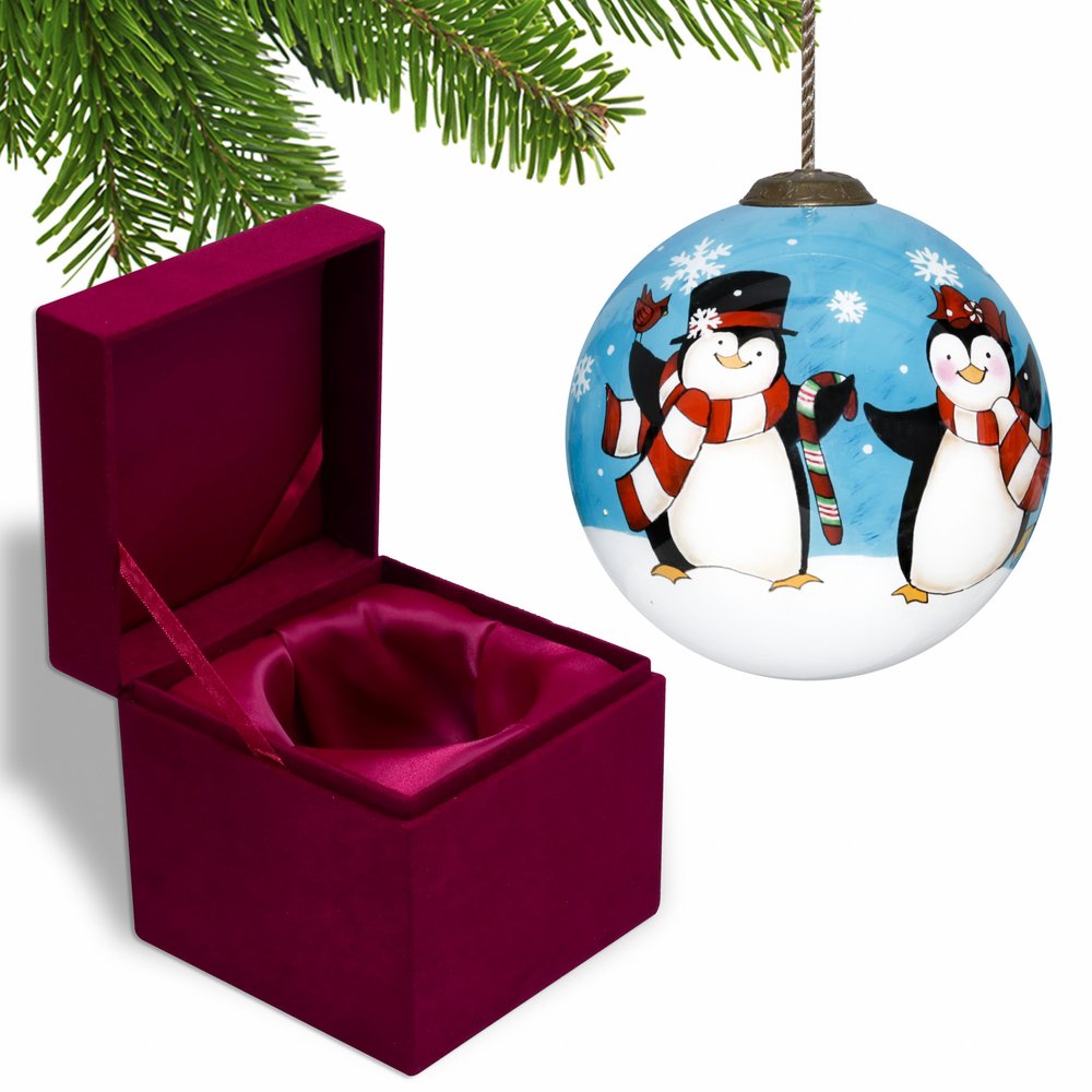 A round glass Christmas ornament with two penguins wearing scarves and hats, set against a blue sky with snowflakes, next to an open velvet gift box.
