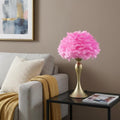 24" Glam Hot Pink Feather and Gold Table Lamp displayed on a dark wood tabletop in a furnished interior against a tan wall.