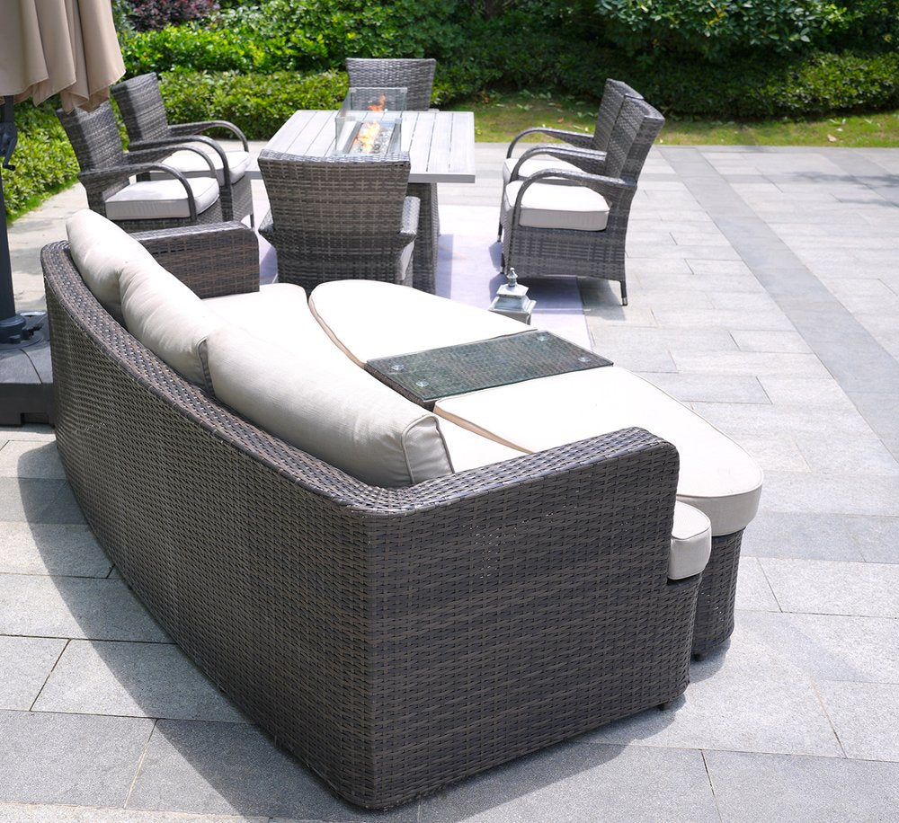 Gray wicker outdoor seating group with fire pit table and chairs on a stone patio with greenery in the background.