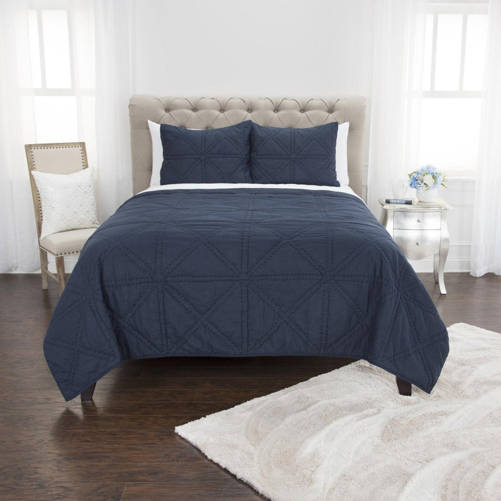 Indigo geometric king quilt set with two pillows on a bed, next to a chair and nightstand in a bedroom.