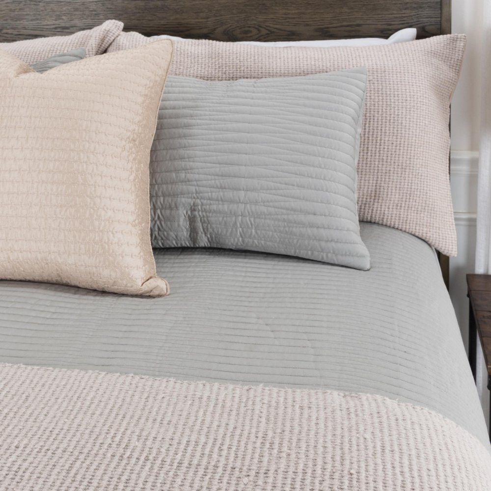 Close-up of a bed with a gray quilted quilt and three pillows in beige and light gray on a wooden headboard.