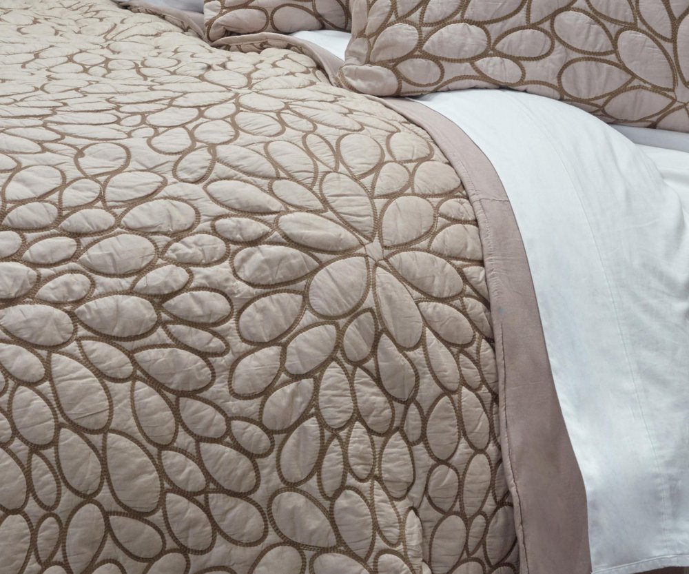 Close-up of a blush floral queen quilt with brown stitching on a bed, featuring a white sheet.