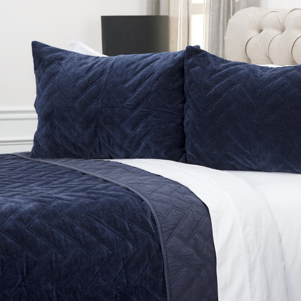 Indigo geometric quilted velvet quilt and pillows on a bed with white sheets and a tufted headboard.