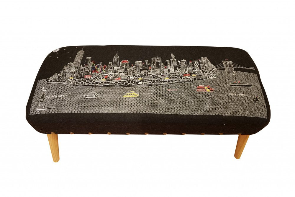 Rectangular ottoman upholstered in gray wool with a black fabric featuring a white and red New York City skyline and river scene.