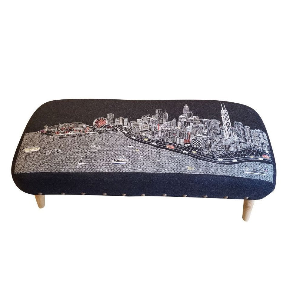 A gray wool ottoman with a Chicago skyline design and wooden legs.