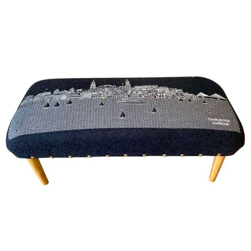 A dark gray upholstered ottoman with a white Charleston Harbour skyline and sailboats design, featuring wooden legs.