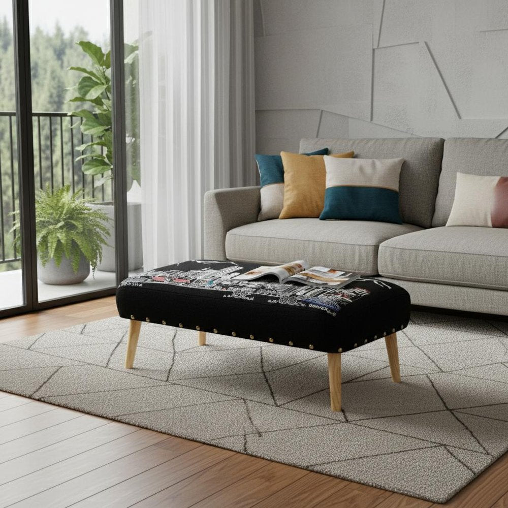 Black ottoman with city skyline embroidery and gold nailhead trim on a geometric rug in a living room.