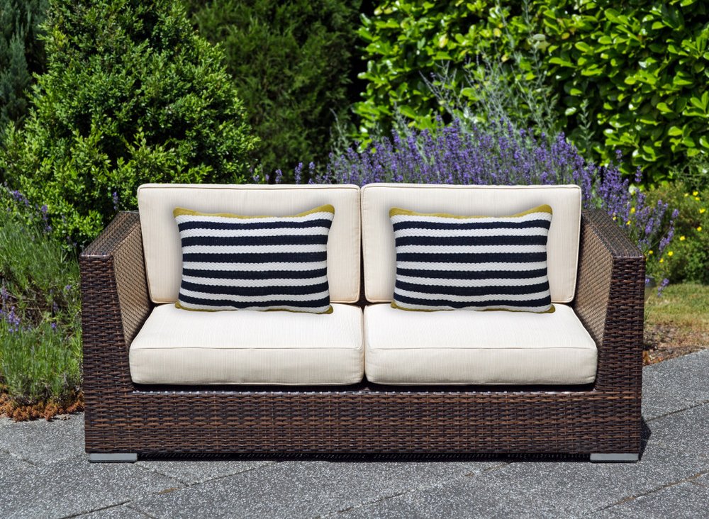 Outdoor loveseat with cream cushions and two black and white striped throw pillows with yellow trim on a stone patio with lush green foliage.