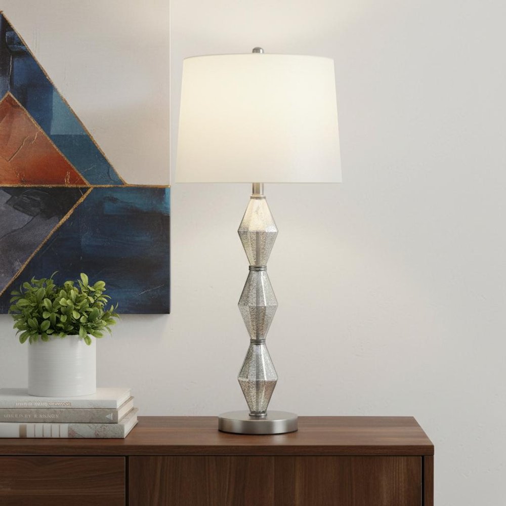 30" Brushed Silver Geo Glass Table Lamp With White Shade displayed on a dark wood tabletop in a furnished interior against an off-white wall.