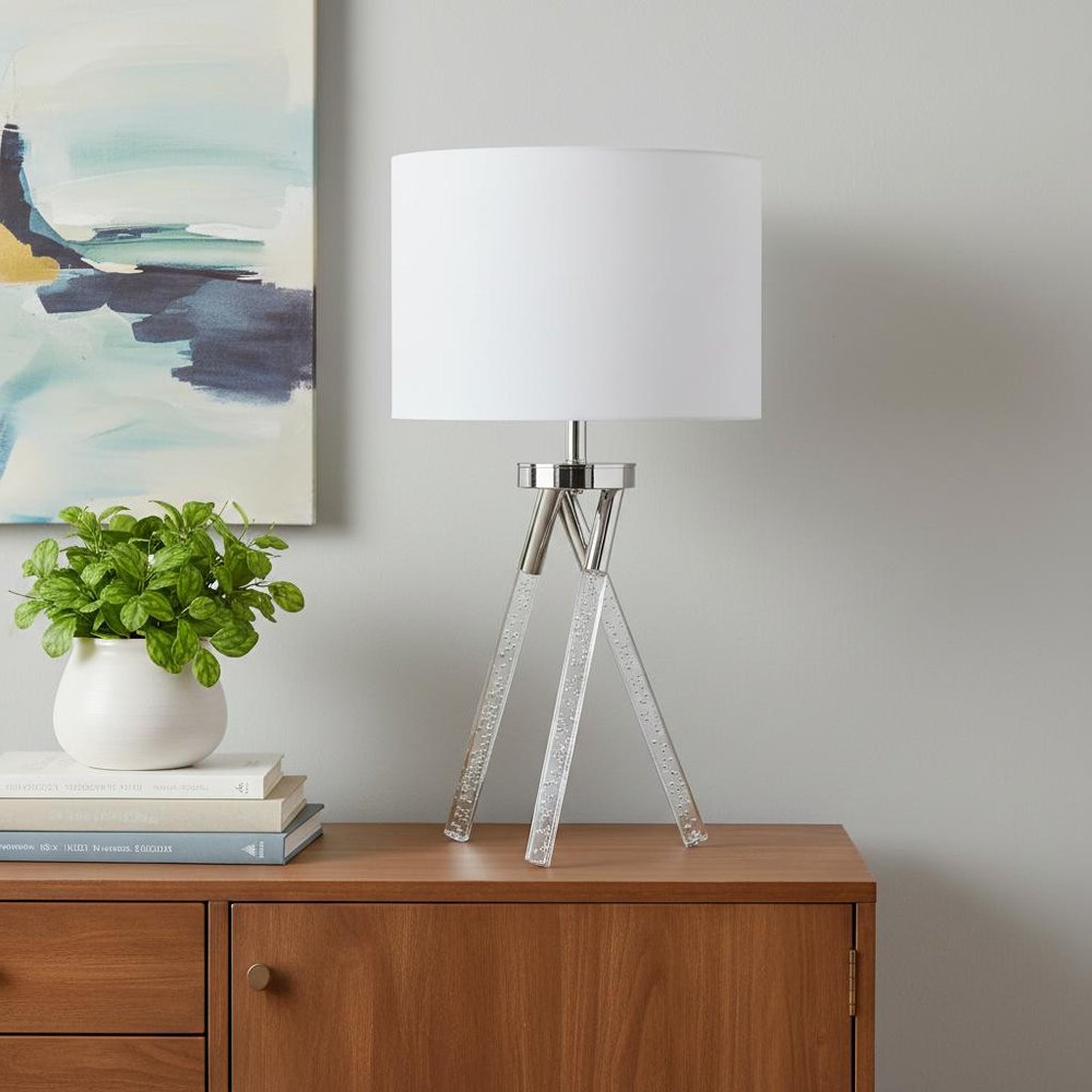 26" Silver and LED Acrylic Tripod Table Lamp With White Shade displayed on a dark wood tabletop in a furnished interior against a light gray wall.