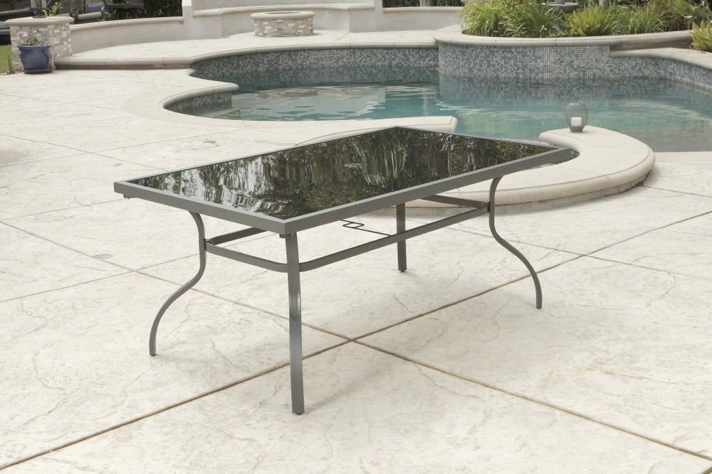 Rectangular glass-top dining table with gray metal frame on a stamped concrete patio next to a swimming pool.