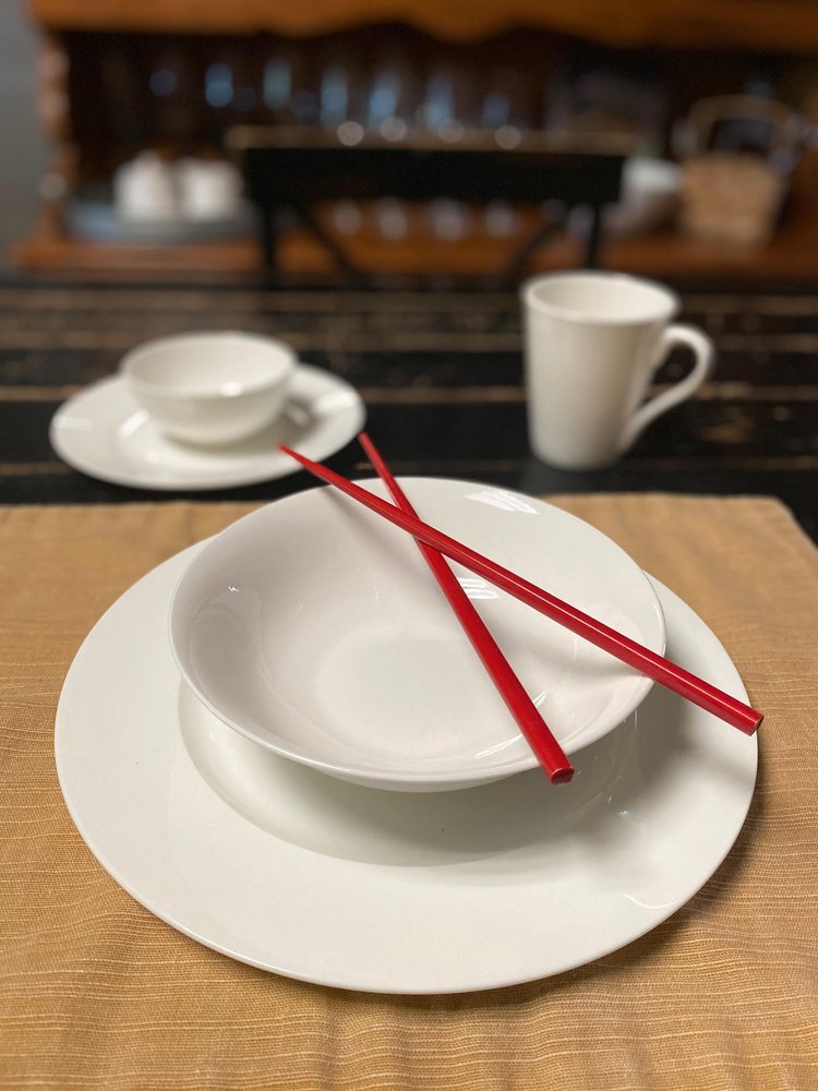 White bone china dinnerware set for four, including plates, bowls, and a mug, with red chopsticks on a tan placemat.