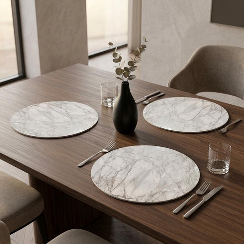 Round marble placemats in silver and gray on a wooden table with place settings and a vase of greenery.