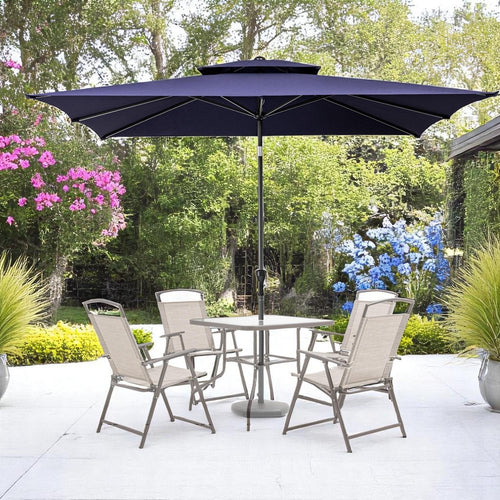 Navy blue rectangular patio umbrella with a tilt feature over a square table and four beige chairs on a white patio with lush green landscaping.