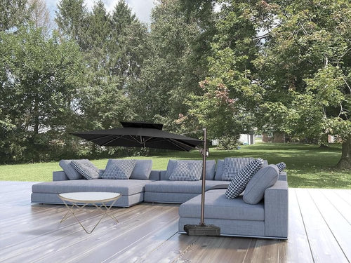 Outdoor patio set with a gray sectional sofa, black cantilever umbrella, and white coffee table on a wooden deck with a green lawn and trees in the background.