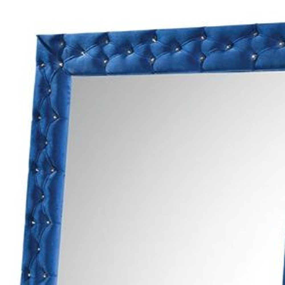Close-up of a blue velvet tufted mirror frame with crystal accents, reflecting a plain background.
