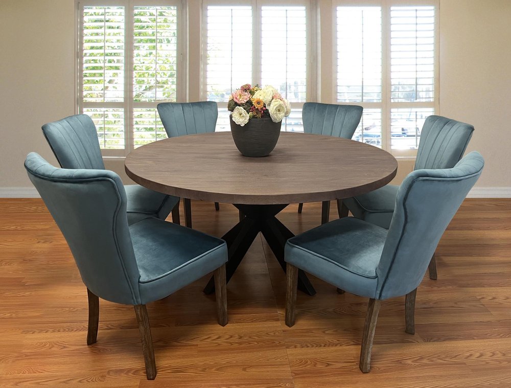Round gray wood dining table with six blue velvet chairs in a room with white shutters and wood floors.