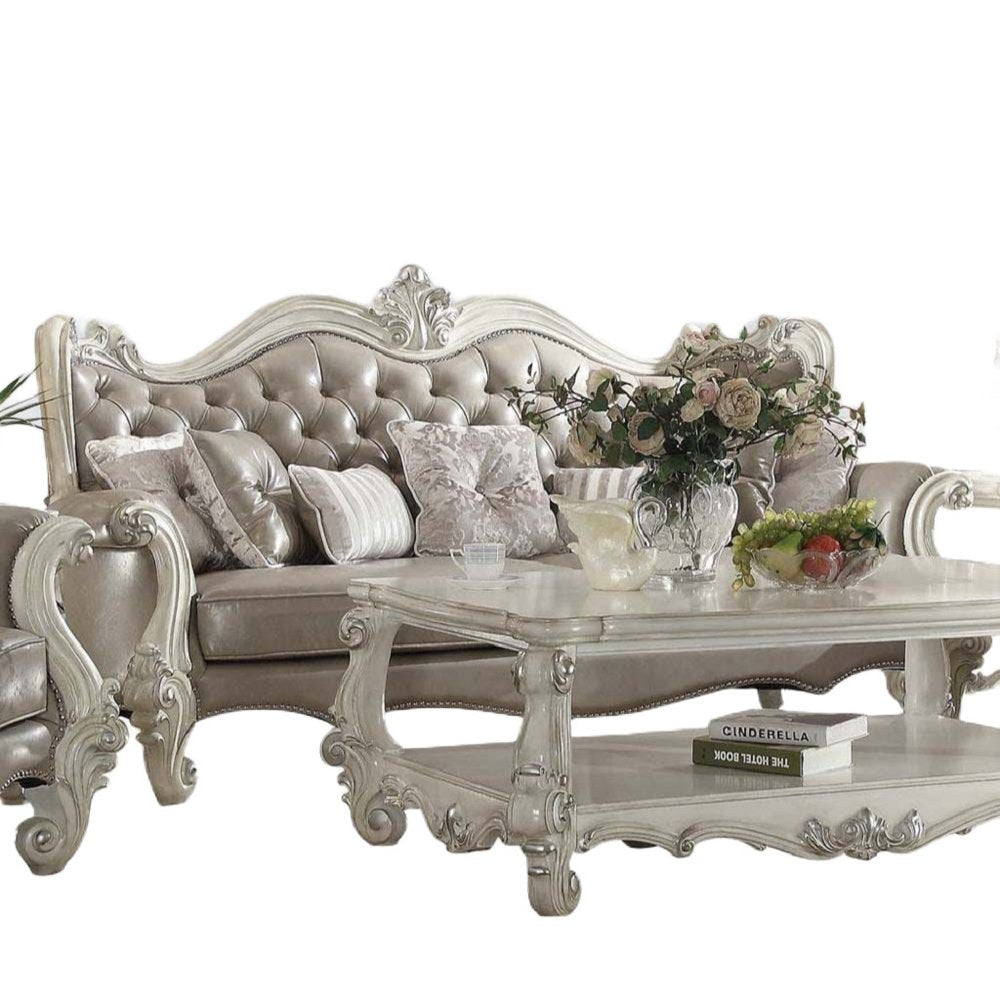 A gray faux leather curved sofa with ornate detailing and toss pillows sits in front of a white background, with a matching coffee table holding a fruit bowl and books.