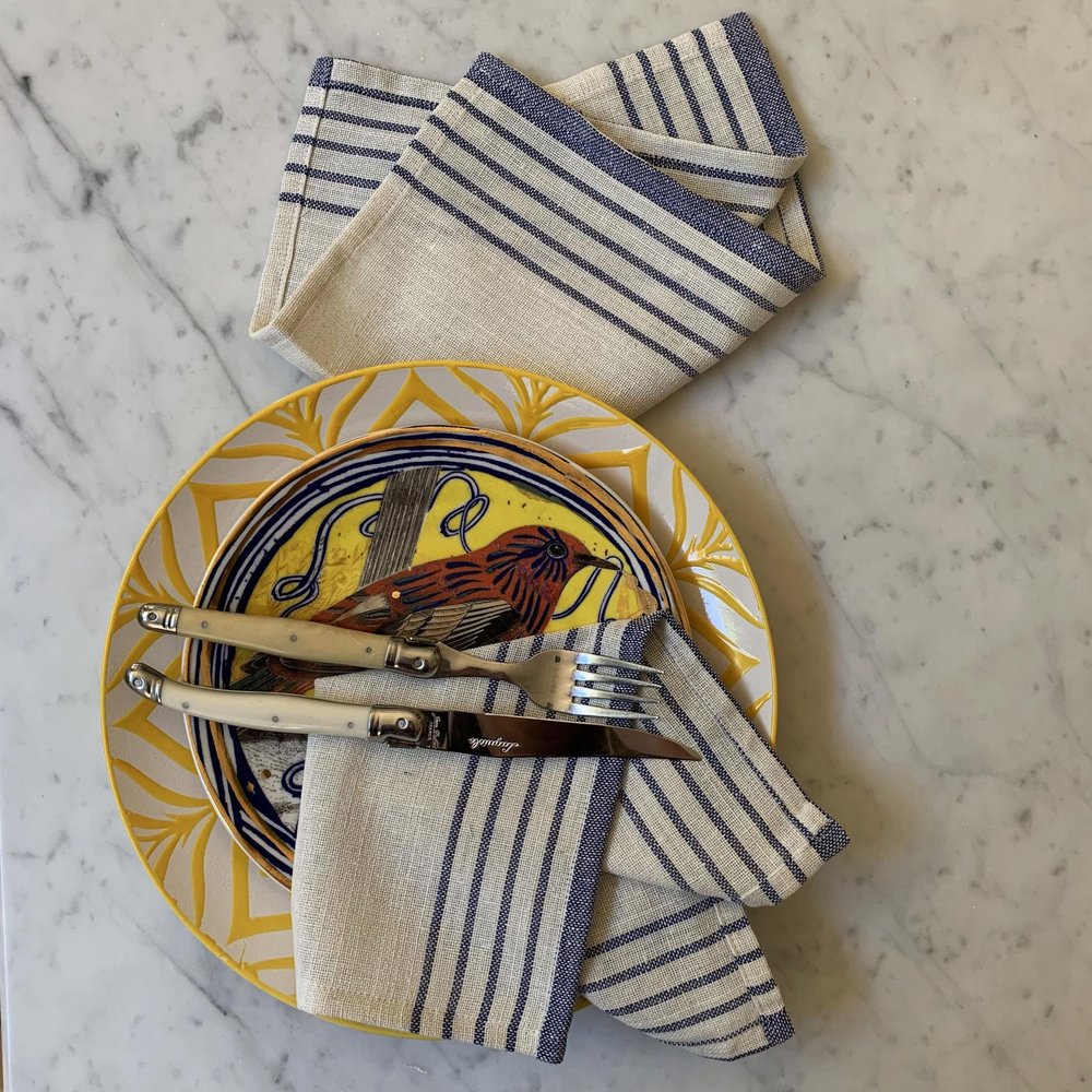 A set of cream and blue striped cotton napkins are folded on a decorative plate with a bird motif, with a knife and fork resting on top.