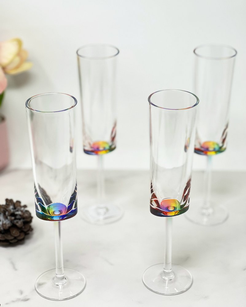 Four clear acrylic stemmed flutes with rainbow accents at the base, arranged on a white marble surface with a pinecone and flowers in the background.