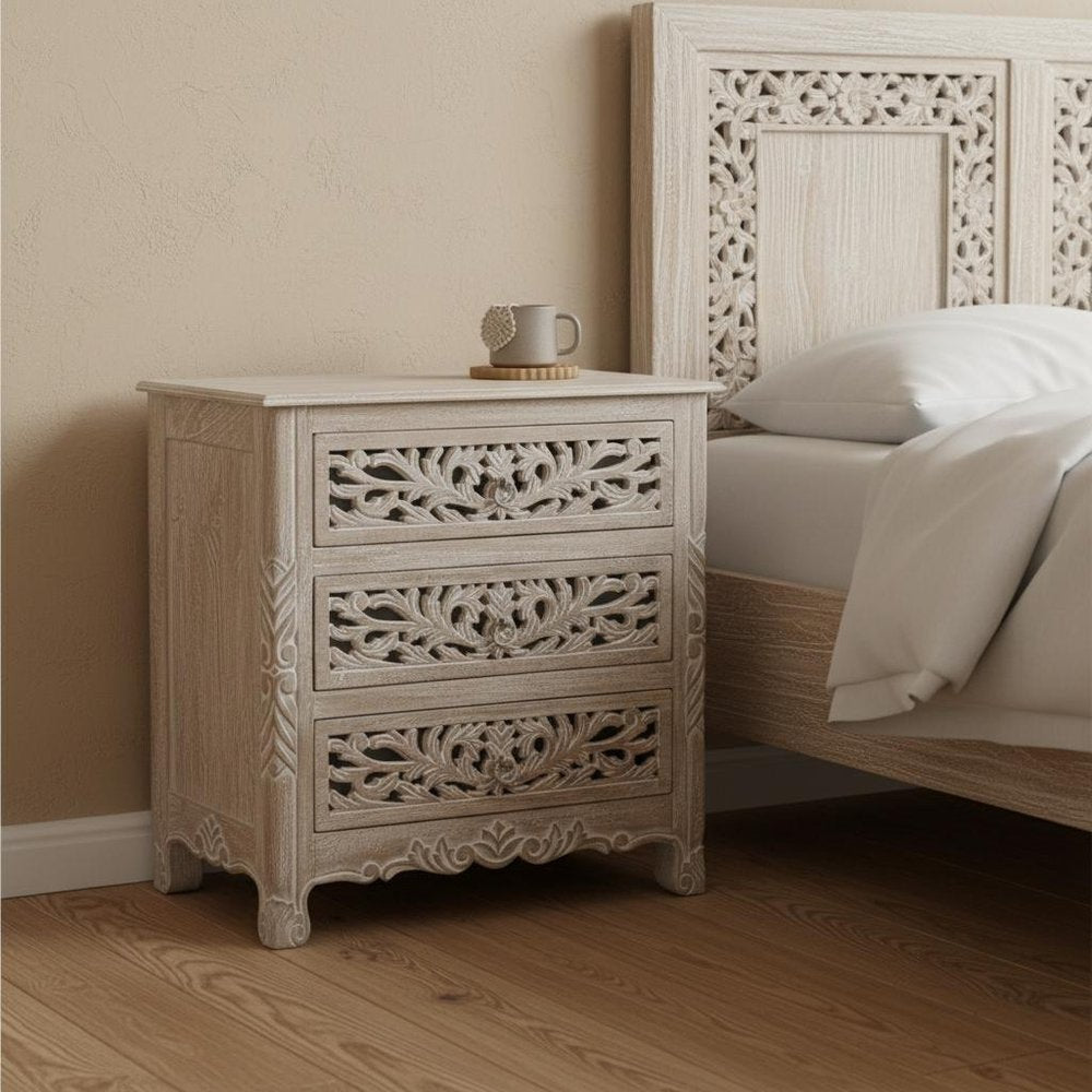 30" Distressed Gray Three Drawer Floral Carved Solid Wood Nightstand styled in a furnished interior with a tan wall on a dark wood floor.