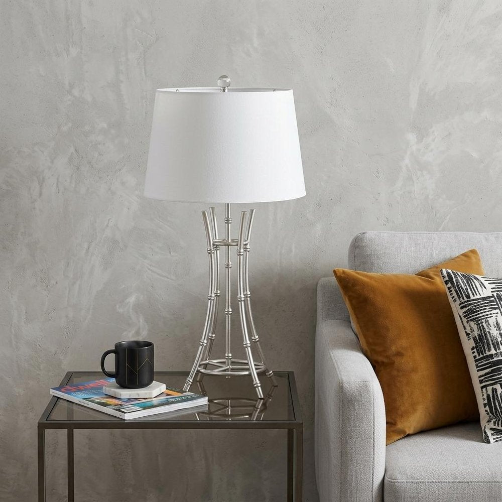 29" Silver Bamboo Design Table Lamp With Off White Drum Shade displayed on a dark wood tabletop in a furnished interior against a light gray wall in off-white.