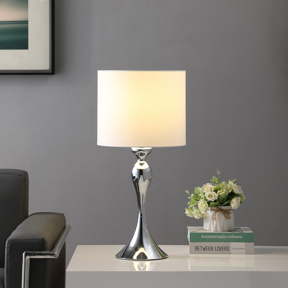 25" Silver Sleek Table Lamp With Off White Drum Shade displayed on a tabletop in a furnished interior against a gray wall in off-white.
