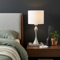 25" Silver Metal Table Lamp With Off White Drum Shade displayed on a dark wood tabletop in a furnished interior against a dark wall.