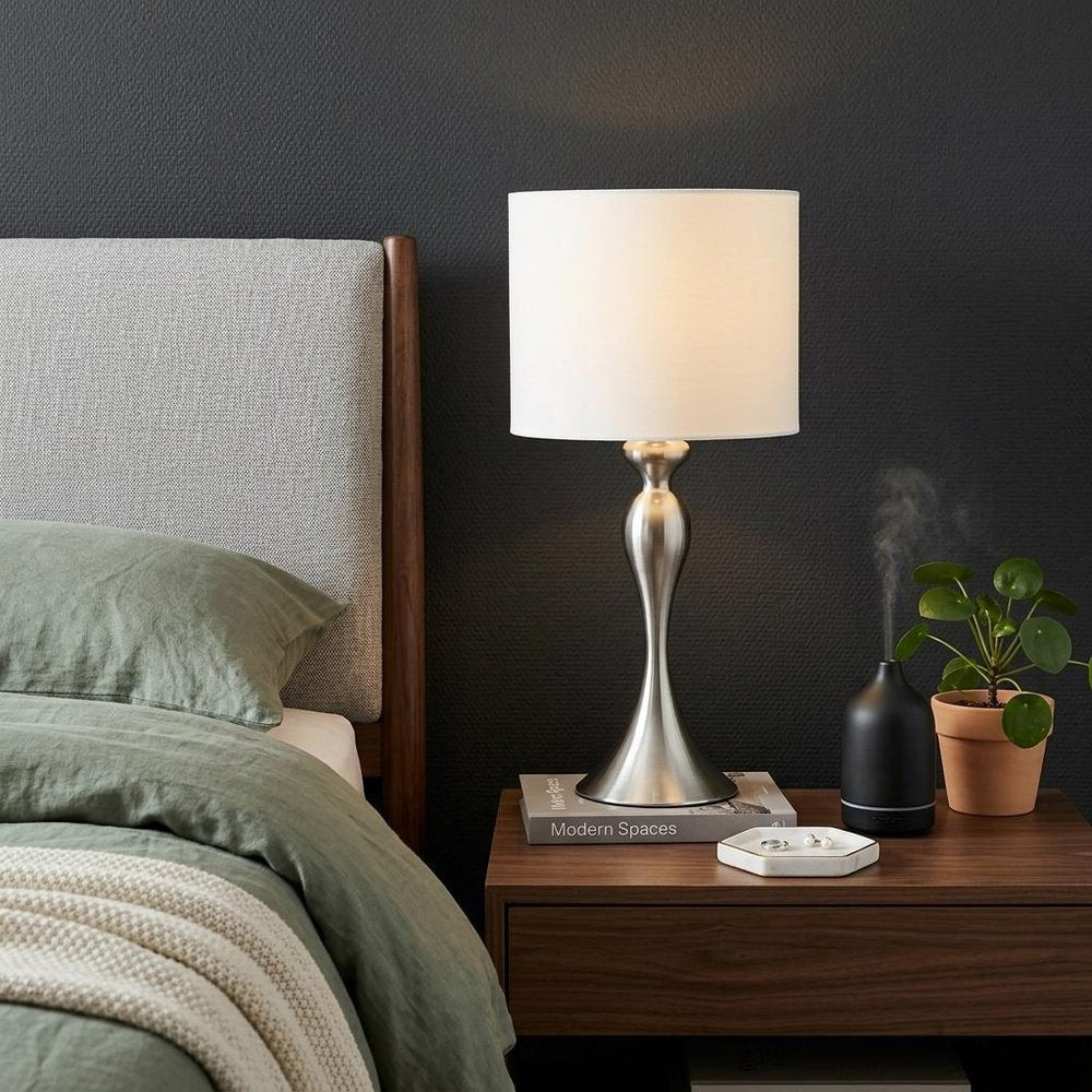 25" Silver Metal Table Lamp With Off White Drum Shade displayed on a dark wood tabletop in a furnished interior against a dark wall.