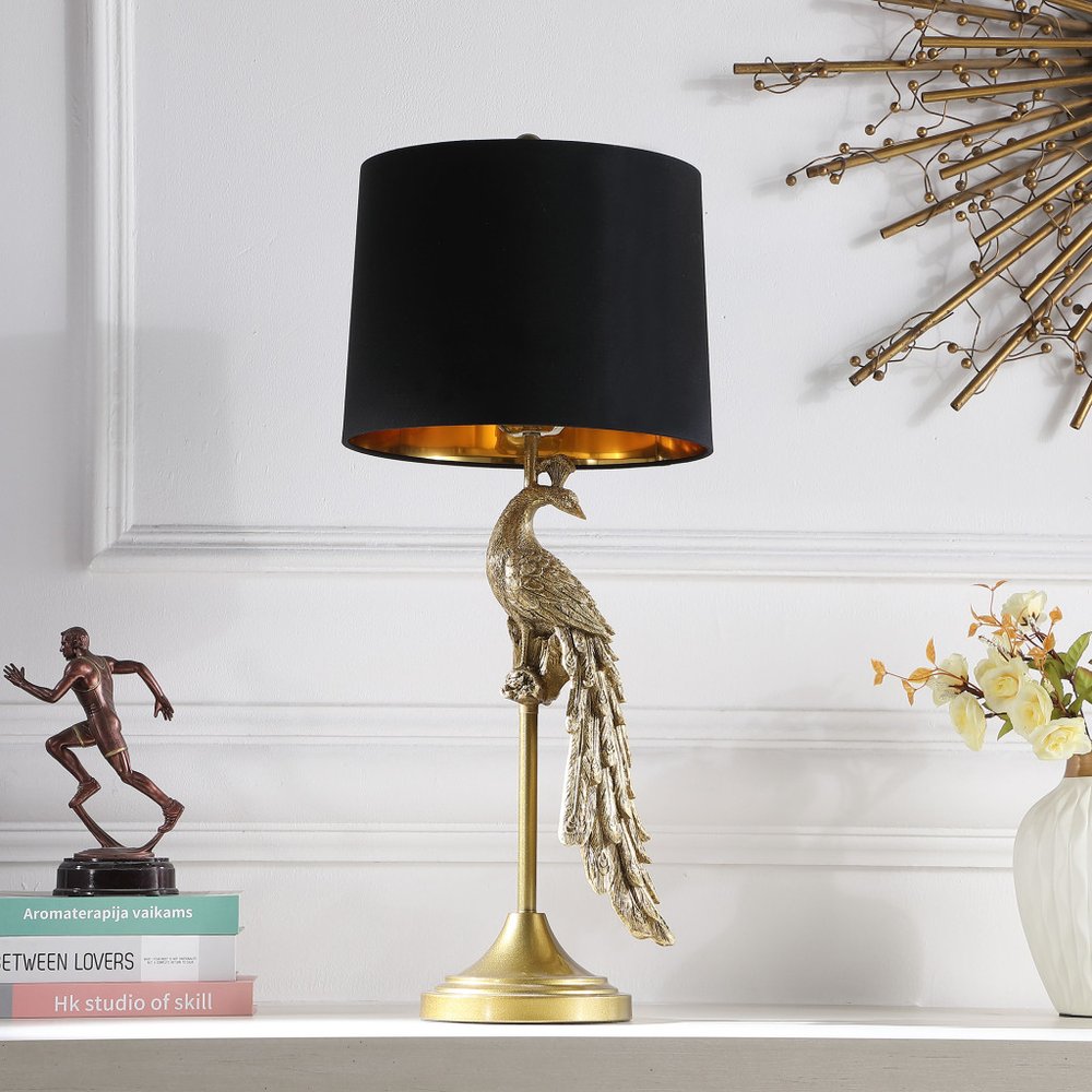 29" Gold Peacock Table Lamp With Black Drum Shade displayed on a tabletop in a furnished interior against an off-white wall.