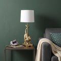 24" Gold Monkey Table Lamp With White Drum Shade displayed on a tabletop in a furnished interior against a dark wall.