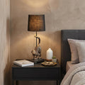 24" Antiqued Gold Monkey Table Lamp With Black Drum Shade displayed on a tabletop in a furnished interior against a tan wall.