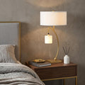 29" Gold Metal Two Light Arched Table Lamp With White Drum Shade displayed on a dark wood tabletop in a furnished interior against a tan wall.