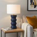28" Navy Blue Geometric Table Lamp With Drum Shade displayed on a dark wood tabletop in a furnished interior against a light gray wall.